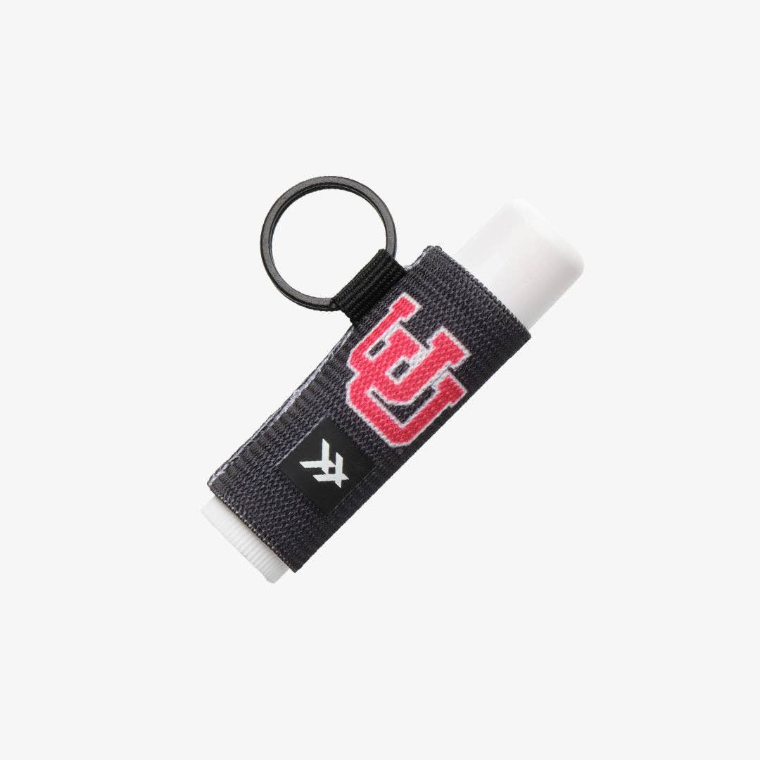 A lip balm tube wrapped in a black textured sleeve featuring a red embroidered 'UC' logo and a white 'U' logo, attached to a black key ring, shown on a light gray background.