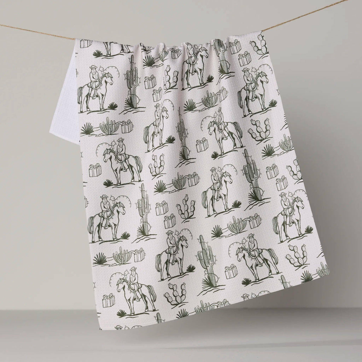 A white fabric tea towel with a black line-art desert cowboy pattern, hanging from a thin string against a neutral gray background.