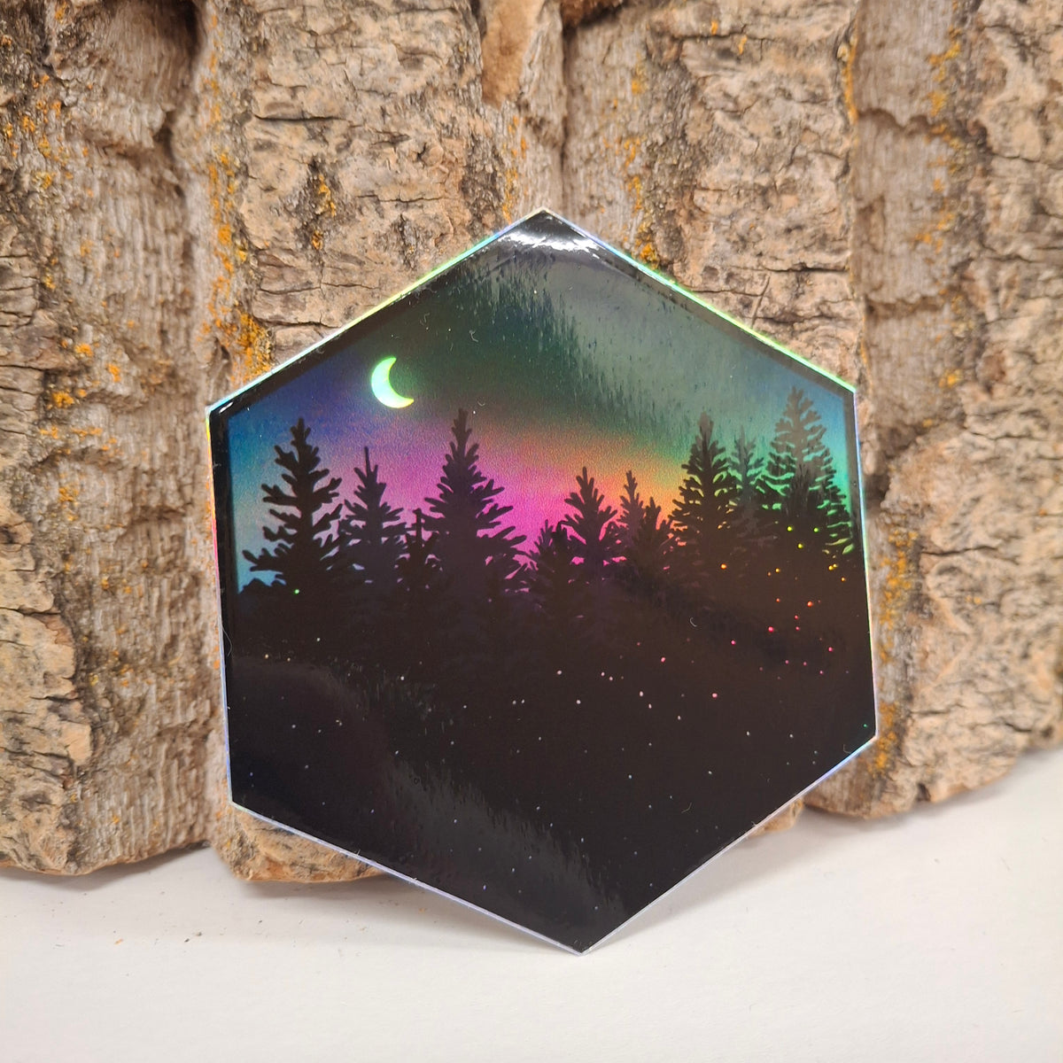 A hexagon sticker featuring the moon surrounded by twinkling stars and majestic pine trees. Holographic with a rainbow sheen.