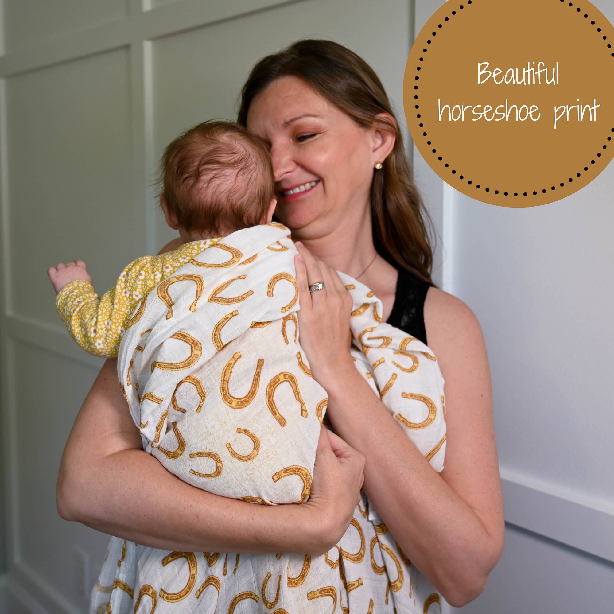 Woman holding a baby wrapped in a blanket with horseshoe pattern against a white wall.