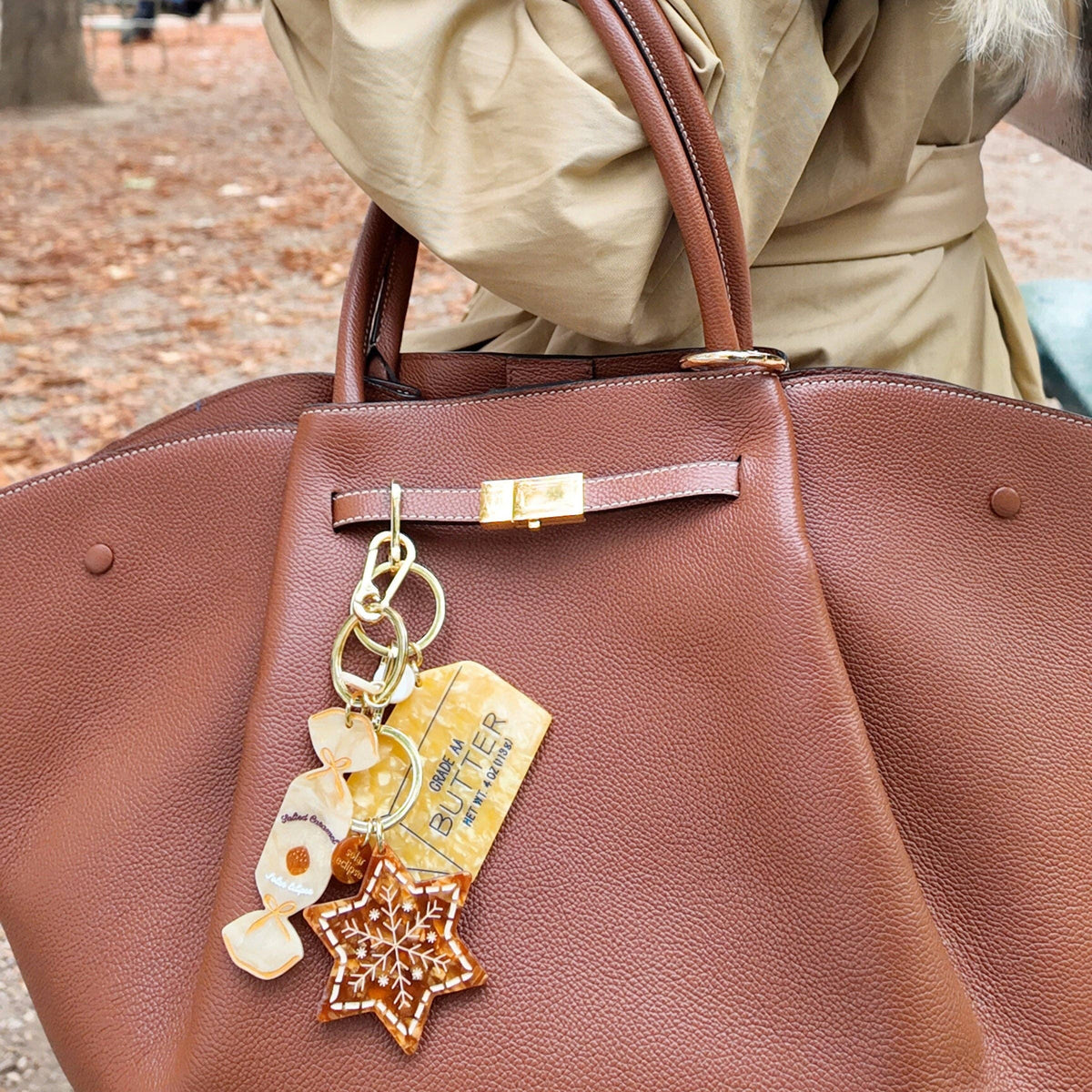 Brown handbag with decorative charms and a person wearing a beige coat in an outdoor setting.