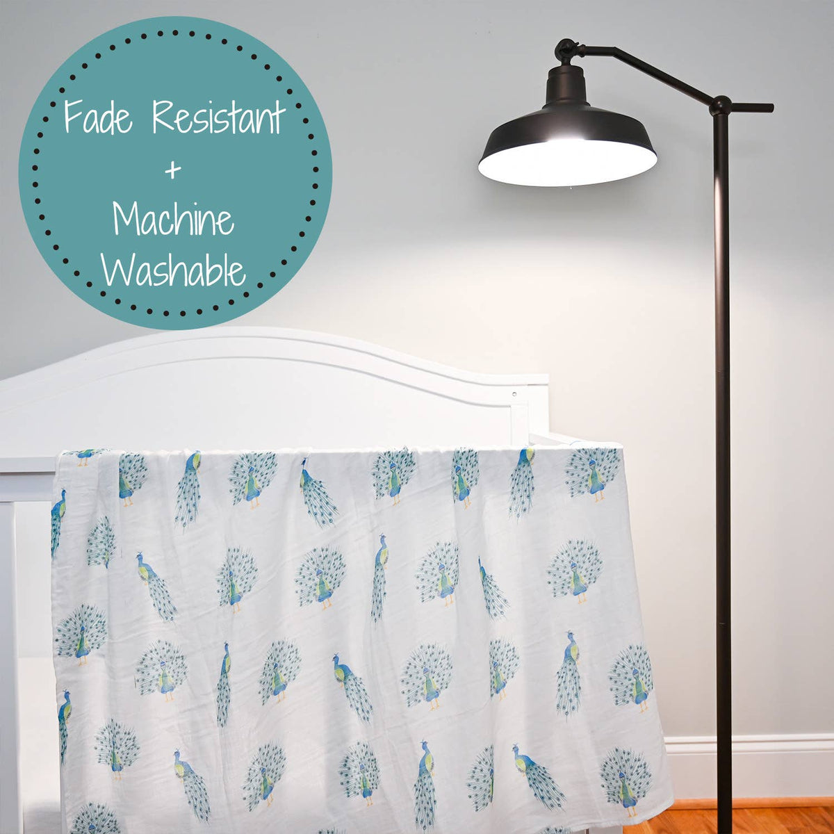 A white baby blanket with a blue peacock pattern draped over a white crib, next to a black floor lamp. A teal circular badge in the top left corner reads 'Fade Resistant + Machine Washable'.
