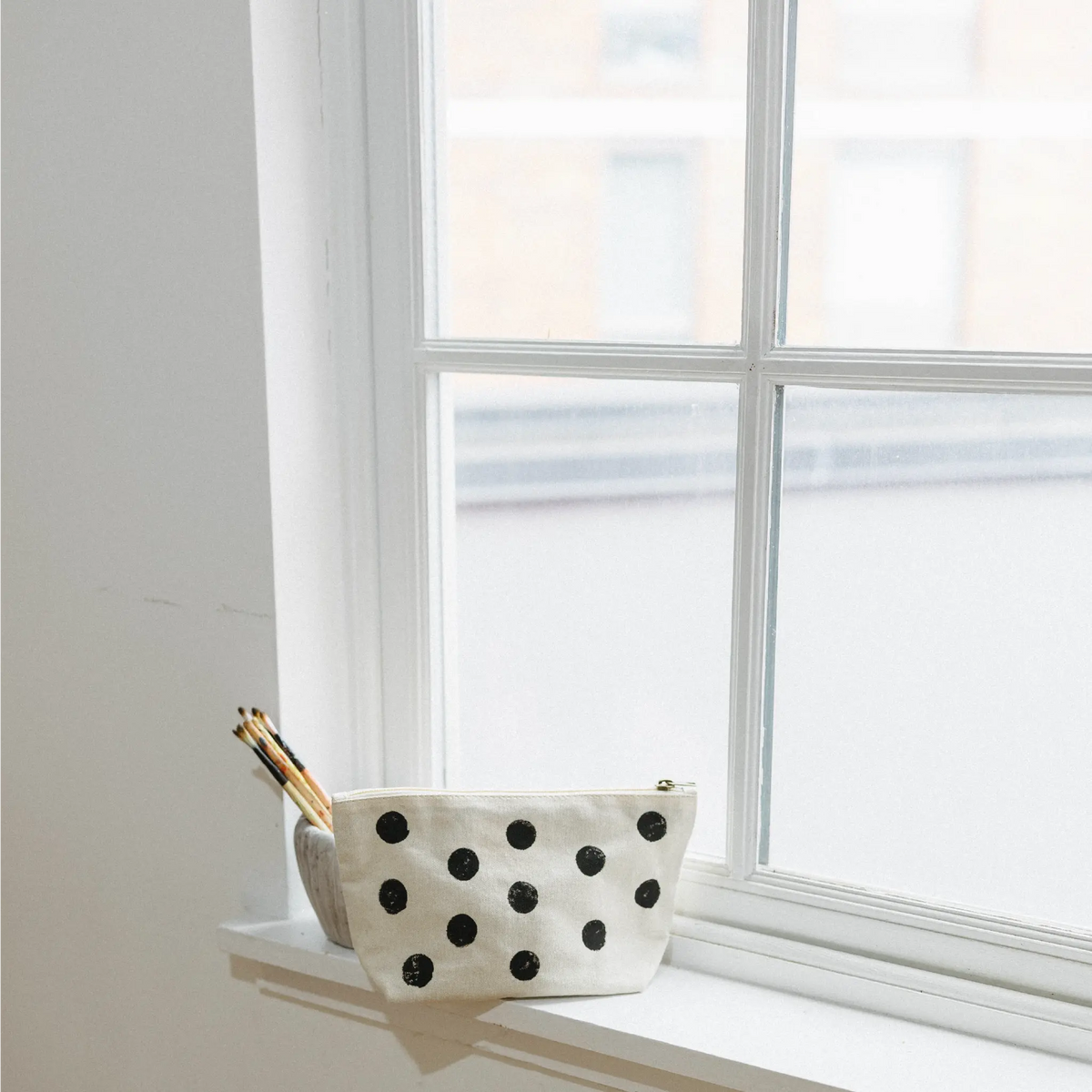 A white canvas zippered pouch with black polka dots sits on a white windowsill next to a bright window, with a few wooden pencils resting inside.