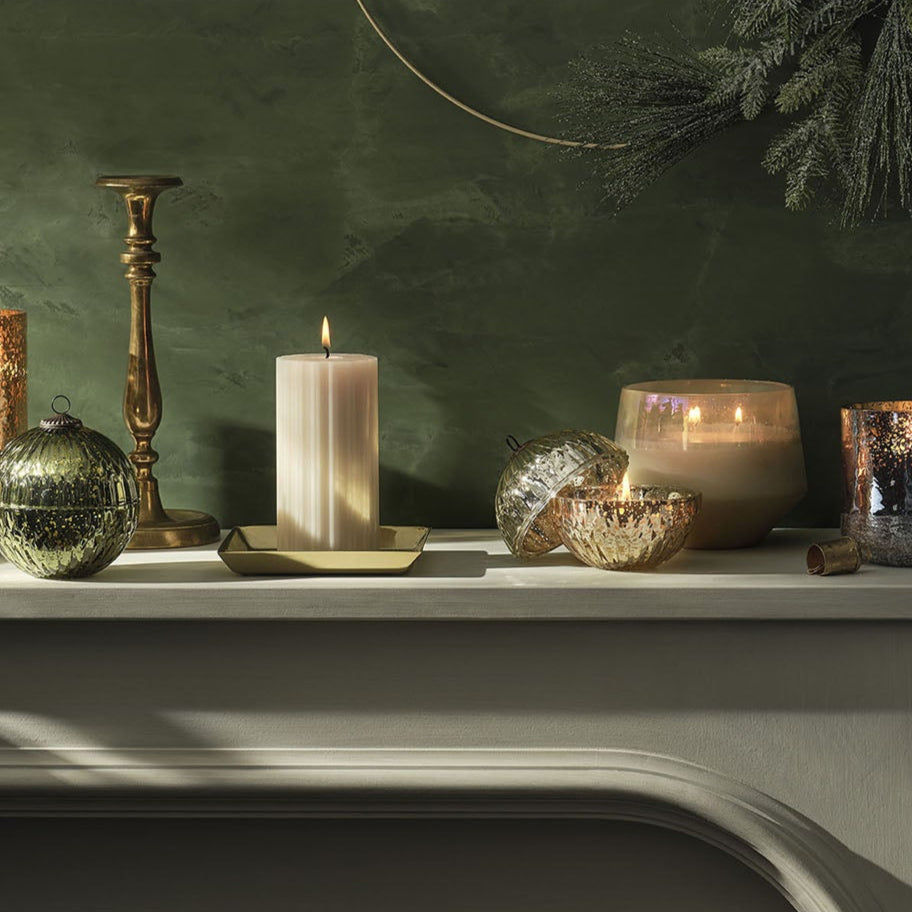 Decorative candles and glass containers on a mantelpiece against a green wall.