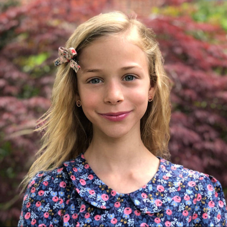 Young girl with a floral head bow and dress standing outdoors with greenery in the background