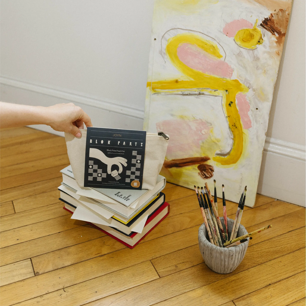 A hand holds a canvas tote bag filled with books on a wooden floor, with an abstract painting and a small pot of paintbrushes in the background.