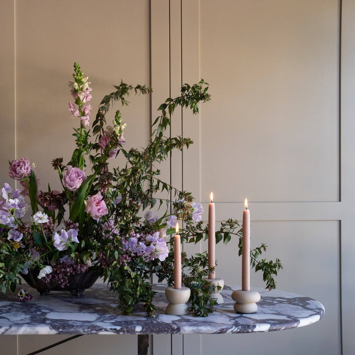 A styled floral arrangement with green foliage and pink and lavender flowers on a marble-topped table, alongside three lit taper candles in matte holders against a beige paneled wall.