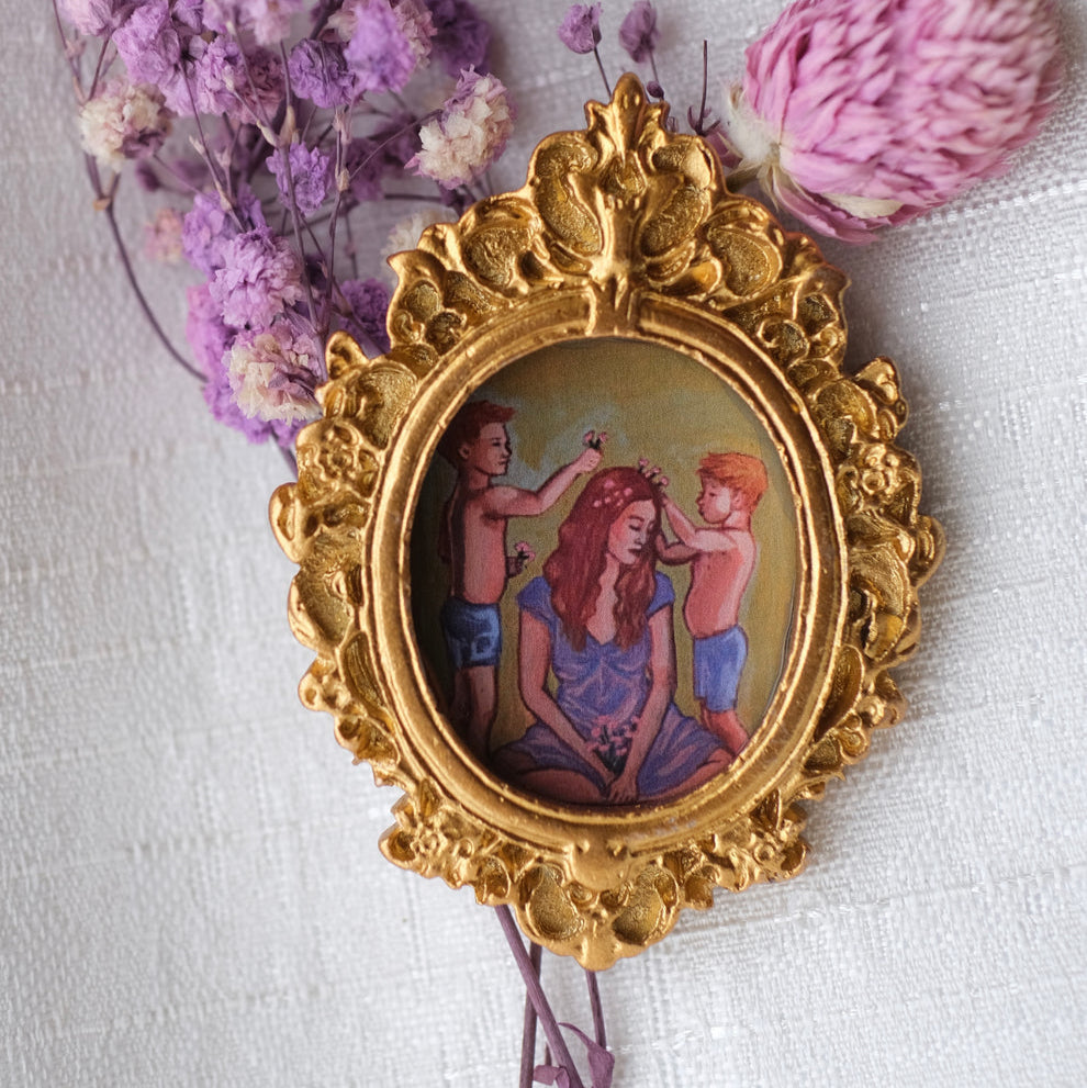 A detailed gold oval frame holds a painting of a woman and two young boys joyfully sitting together, they boys are playing with the mothers hair. The artwork is set against a pale yellow background and is surrounded by sprigs of dried flowers on a textured fabric.