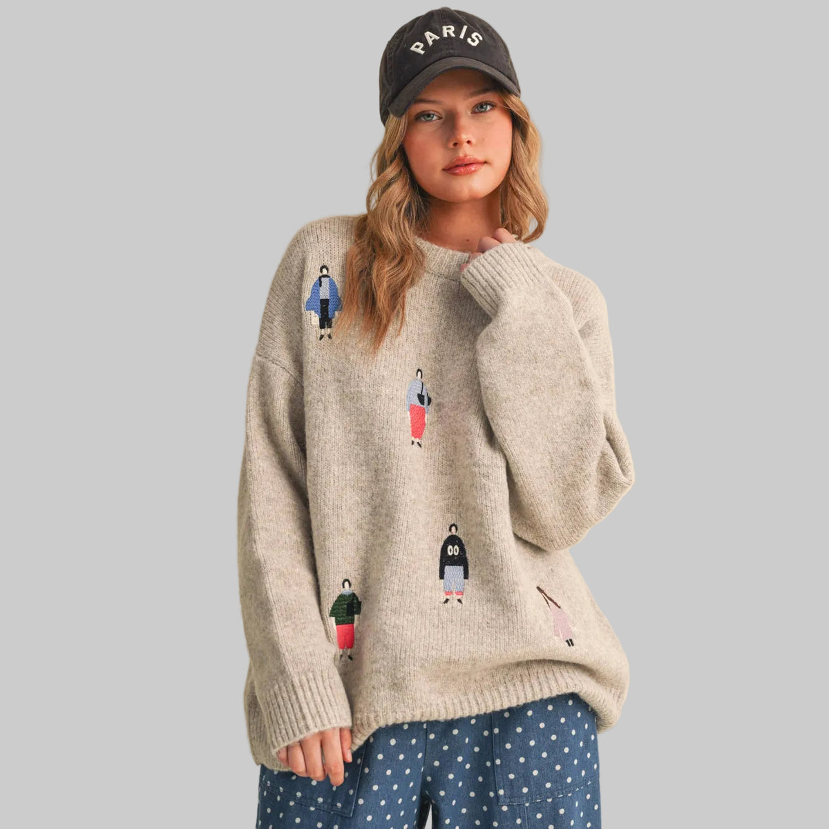 A woman wearing a beige knit sweater with small colorful women dressed in fun clothing patches, paired with gray polka-dot pants, against a plain beige studio background.