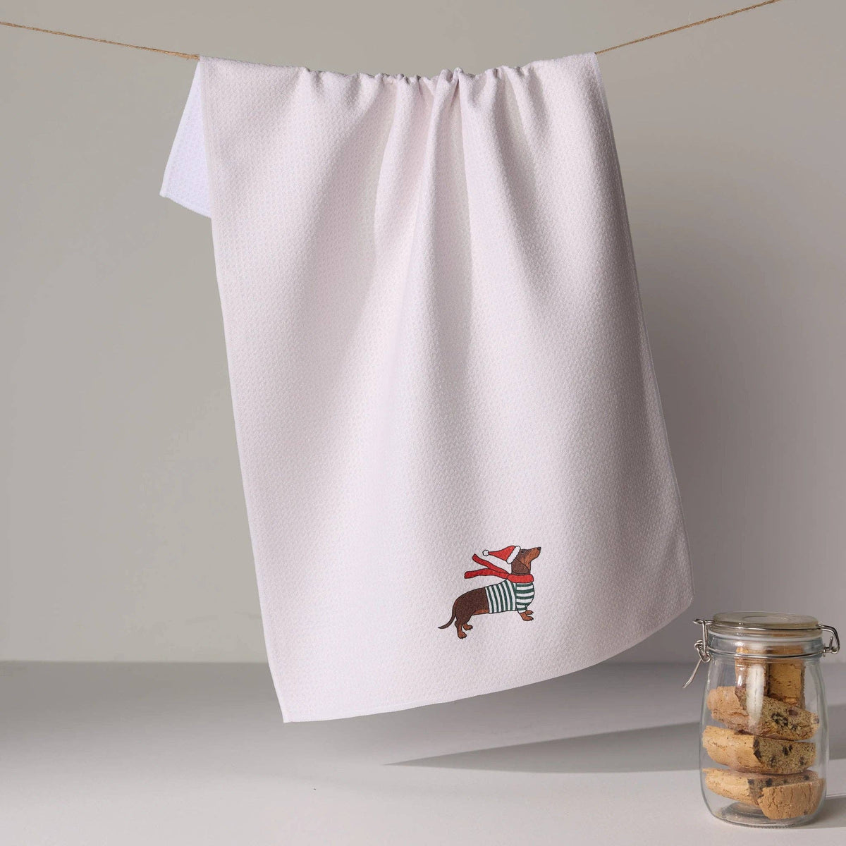 A white kitchen tea towel hangs from a string on a wall, featuring a small dachshund wearing a red Santa hat and striped sweater near the bottom. A glass jar of cookies sits on the surface below.