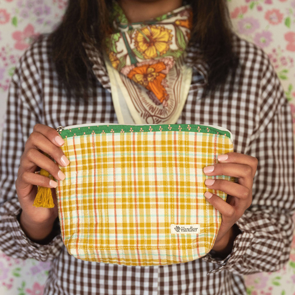 Person holding a yellow and green checkered pouch with a floral-patterned scarf around their neck against a floral background.