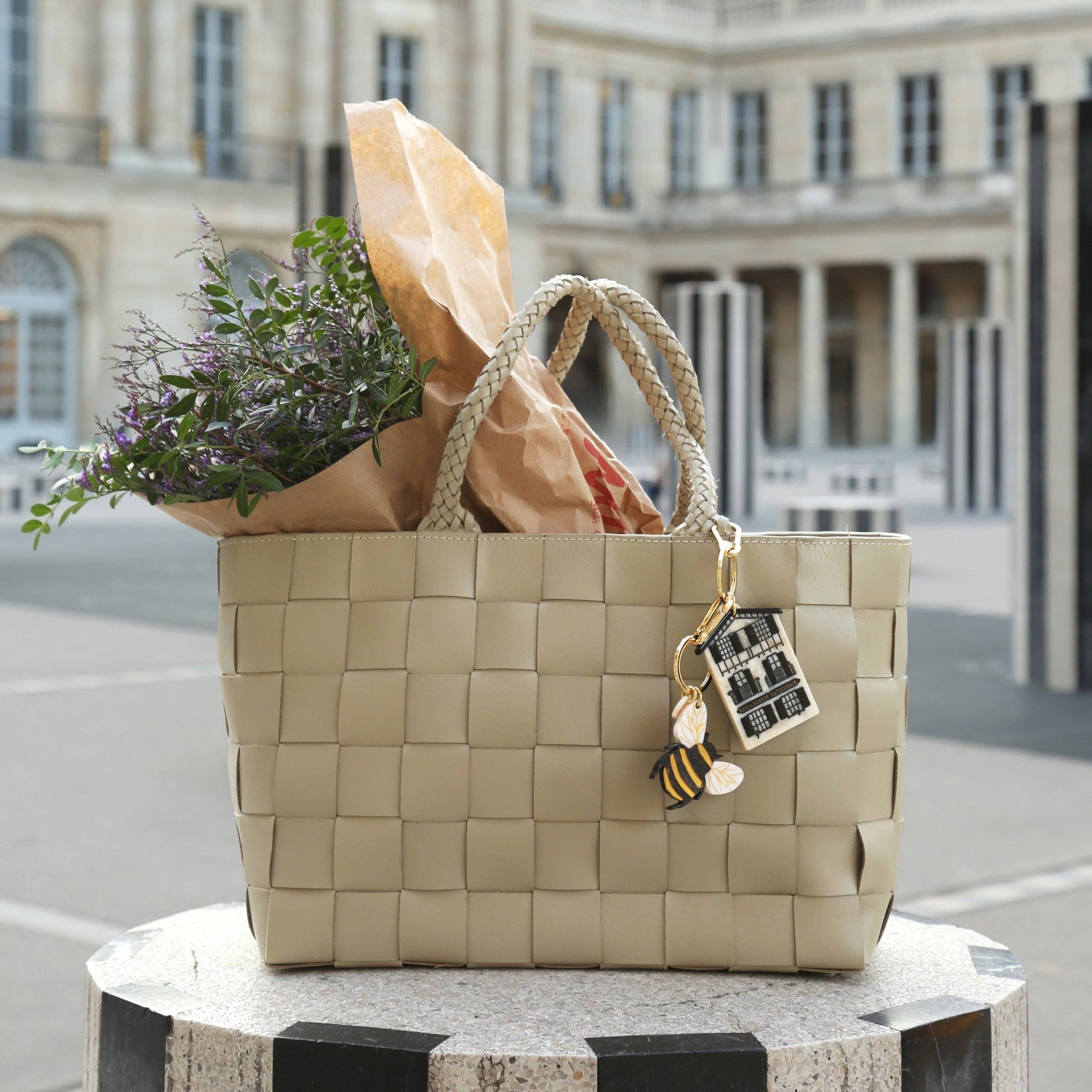 Woven beige bag with flowers and a paper bag on a pedestal in front of an architectural building.