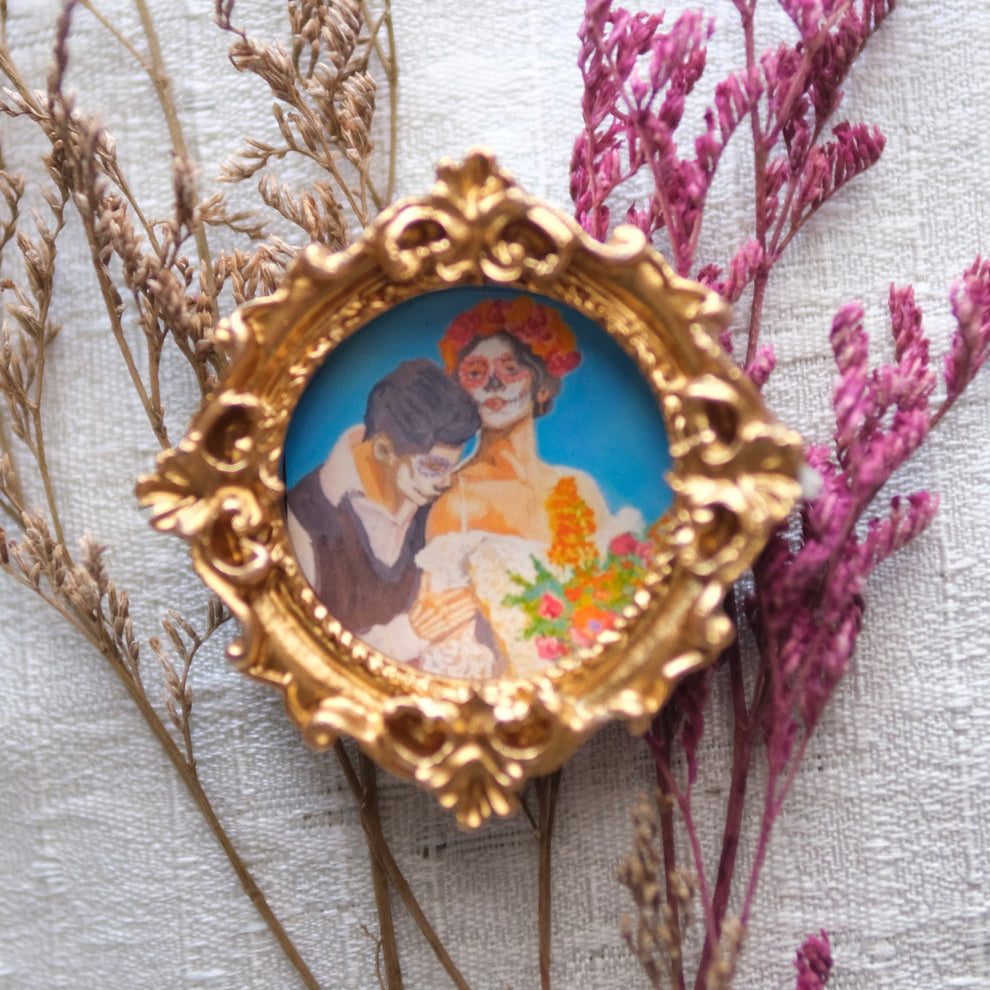 A small ornate gold frame contains a painted scene of a couple embracing against a bright blue sky, the woman with Día de los Muertos skull makeup and a vibrant flower crown, holding a bouquet of marigolds while the man rests his head on her shoulder. The frame is surrounded by delicate dried beige and purple branches on a soft white fabric background.