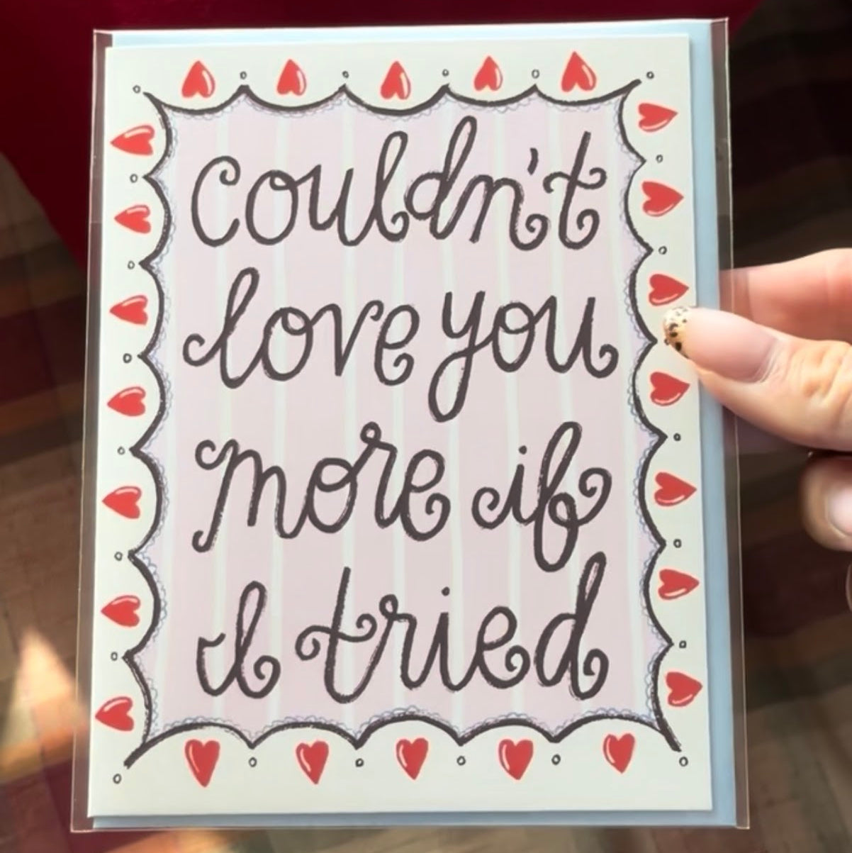 A heartfelt greeting card in a clear plastic sleeve displays the playful handwritten message "Couldn't love you more if I tried" in elegant black cursive lettering, centered on a light pink background with subtle vertical stripes.
The card is bordered by a scalloped frame adorned with numerous small red hearts and tiny white dots, creating a sweet, romantic, and affectionate design perfect for expressing deep love or appreciation.