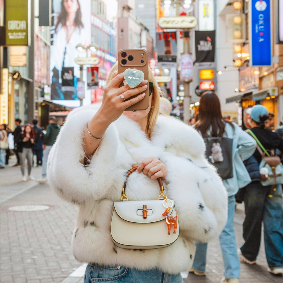Person in a white fur coat taking a selfie in a busy city street with neon signs and pedestrians.