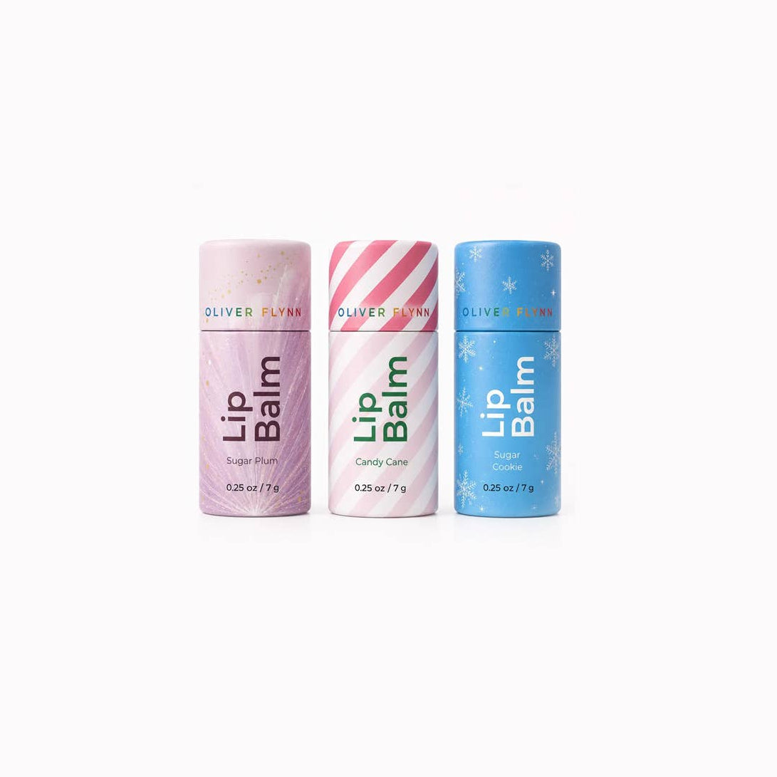 Three Oliver Flynn Lip Balm tubes lined up on a white background: a pink Sugar Elm tube, a white and pink striped Candy Care tube, and a blue Snowflake tube.