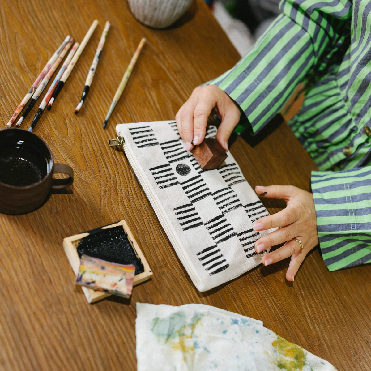 A person wearing a green and gray striped shirt is shown from the side, using a wooden stamp to print a black geometric pattern onto a white canvas. The scene is set on a wooden table with traditional calligraphy tools, including brushes, ink, and a cup.