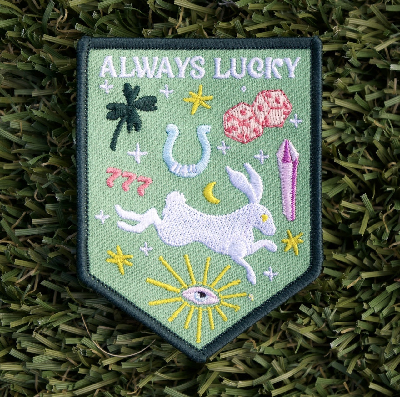 Iron-on patch with a green background, and embroidered rabbit, horseshoe, diamond, four-leaf clover, dice, and the white words "Always Lucky".