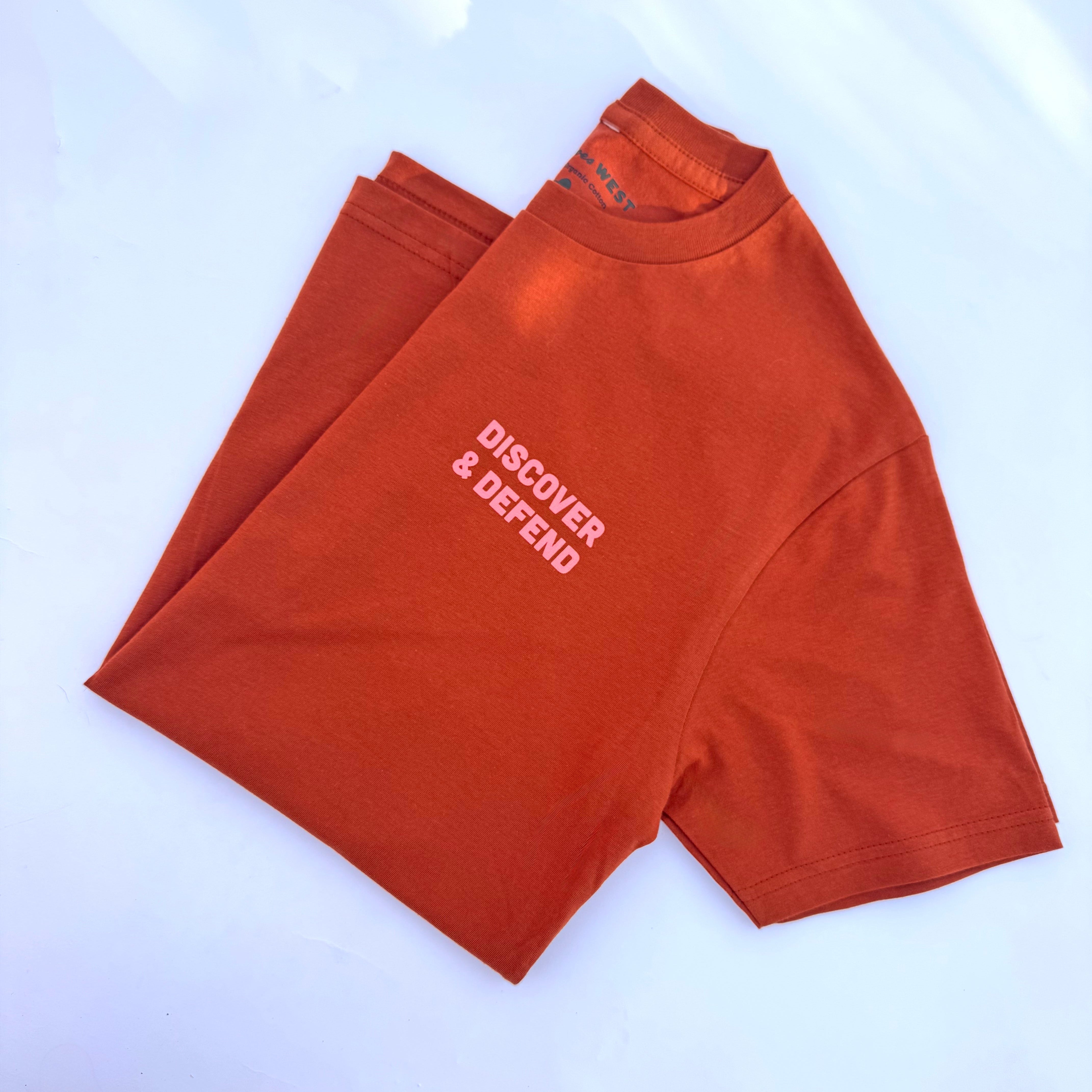 Hand-drawn graphics in faded blush and vibrant pink pop against the fabric's warm, earthy orange tone. A simple, bold front states the mission, "Discover & Defend." The back features a checklist of federally-managed parks in Utah, surrounded by desert flora, fauna, and landmarks.