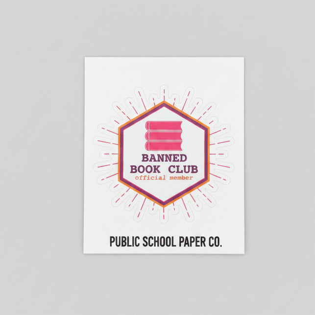 A small white rectangular card or sticker with a red and pink geometric logo reading 'Banned Book Club' and 'Public School Paper Co.' placed on a wooden tabletop.