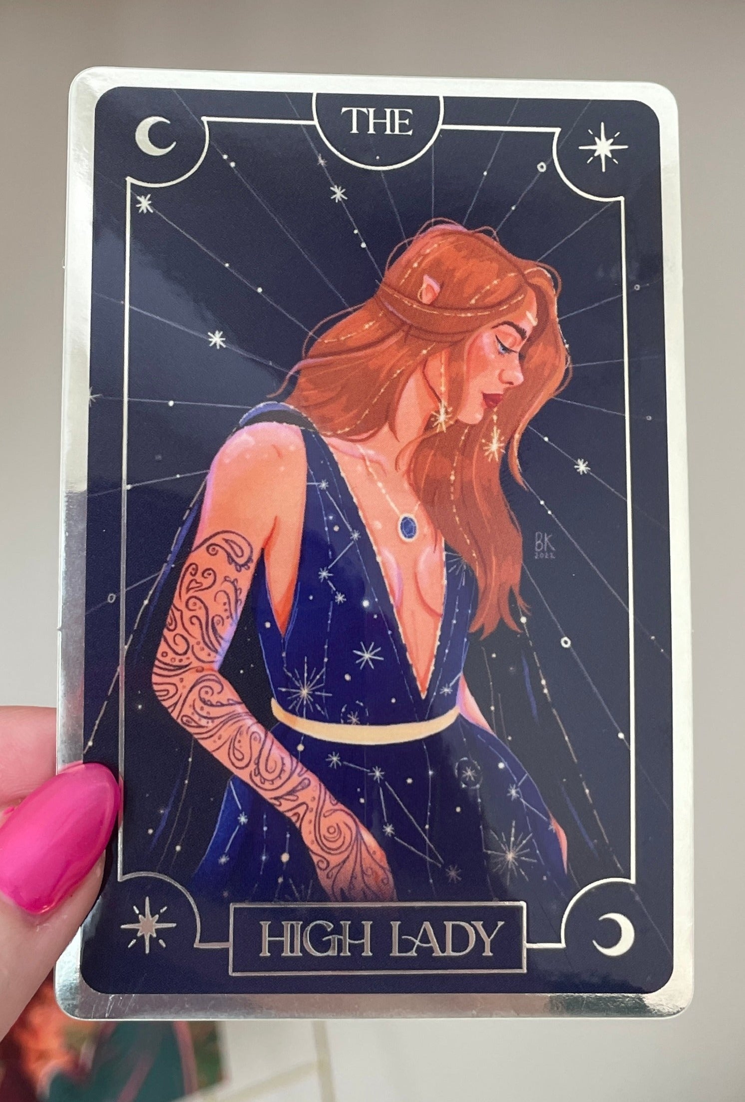Feyre (The High Lady) Sticker with mirrored foil effects. The sticker resembles a tarot card with "HIGH LADY" in print at the bottom. Feyre is a woman with light brown hair and golden accents and she is wearing long earrings that shine like stars. She is wearing a sleeveless dress with a cape and plunging neckline and the dress is adorned with constellations.