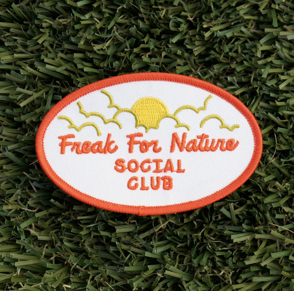 Oval-shaped iron-on embroidered patch with a white background and red words that say "Freak for nature social club" and a yellow sun and clouds.