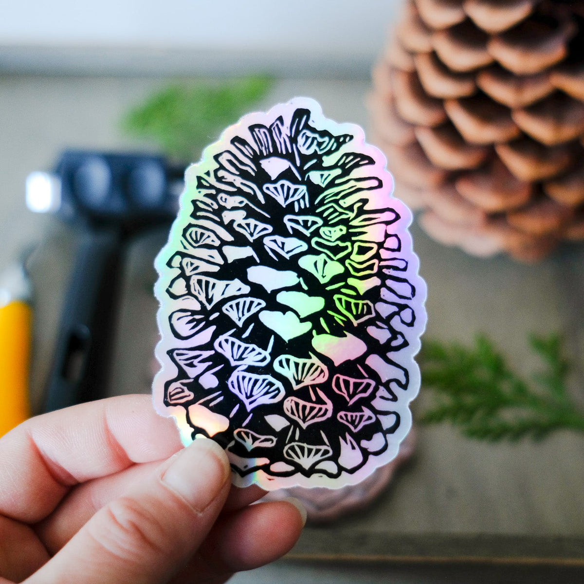 Holographic shimmery sticker with the black detailing of a pinecone. Pictured in a woman's hand with a real pinecone in the back ground.