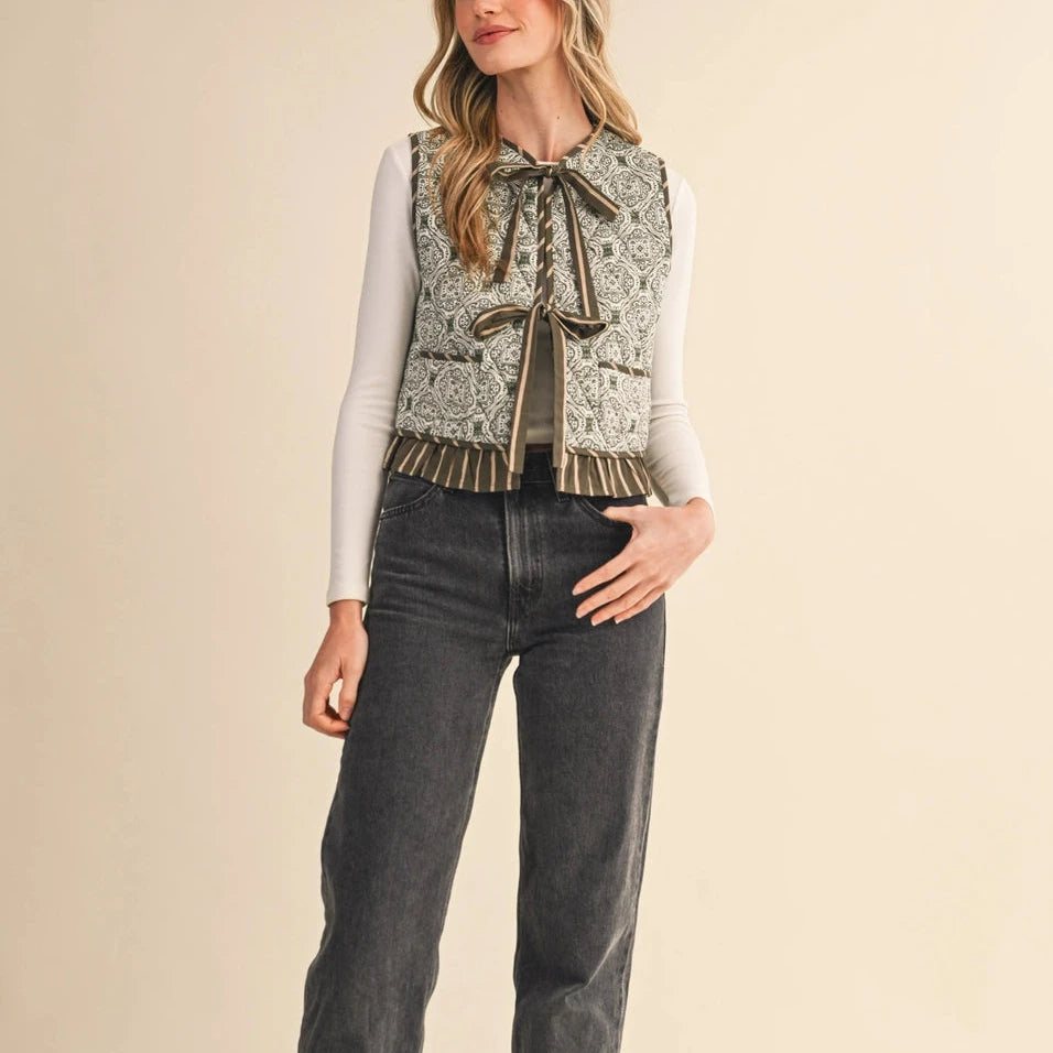 Elevate your layering game with this charming olive green floral quilted vest. Crafted from soft quilted fabric adorned with delicate floral patterns, it features a front-tie closure that lets you customize the fit to your comfort. The ruffle trim adds a touch of feminine elegance to any outfit. Perfect for layering over your favorite tees or dresses, this versatile piece brings warmth and style to your everyday wardrobe.