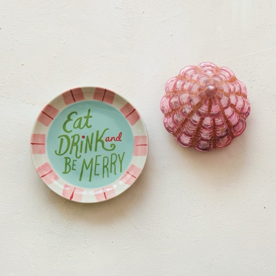 A small round decorative plate with the phrase 'Eat DRINK and BE MERRY' in green lettering on a mint background, surrounded by pink and white stripes. A pink seashell sits beside it on a plain off-white surface.
