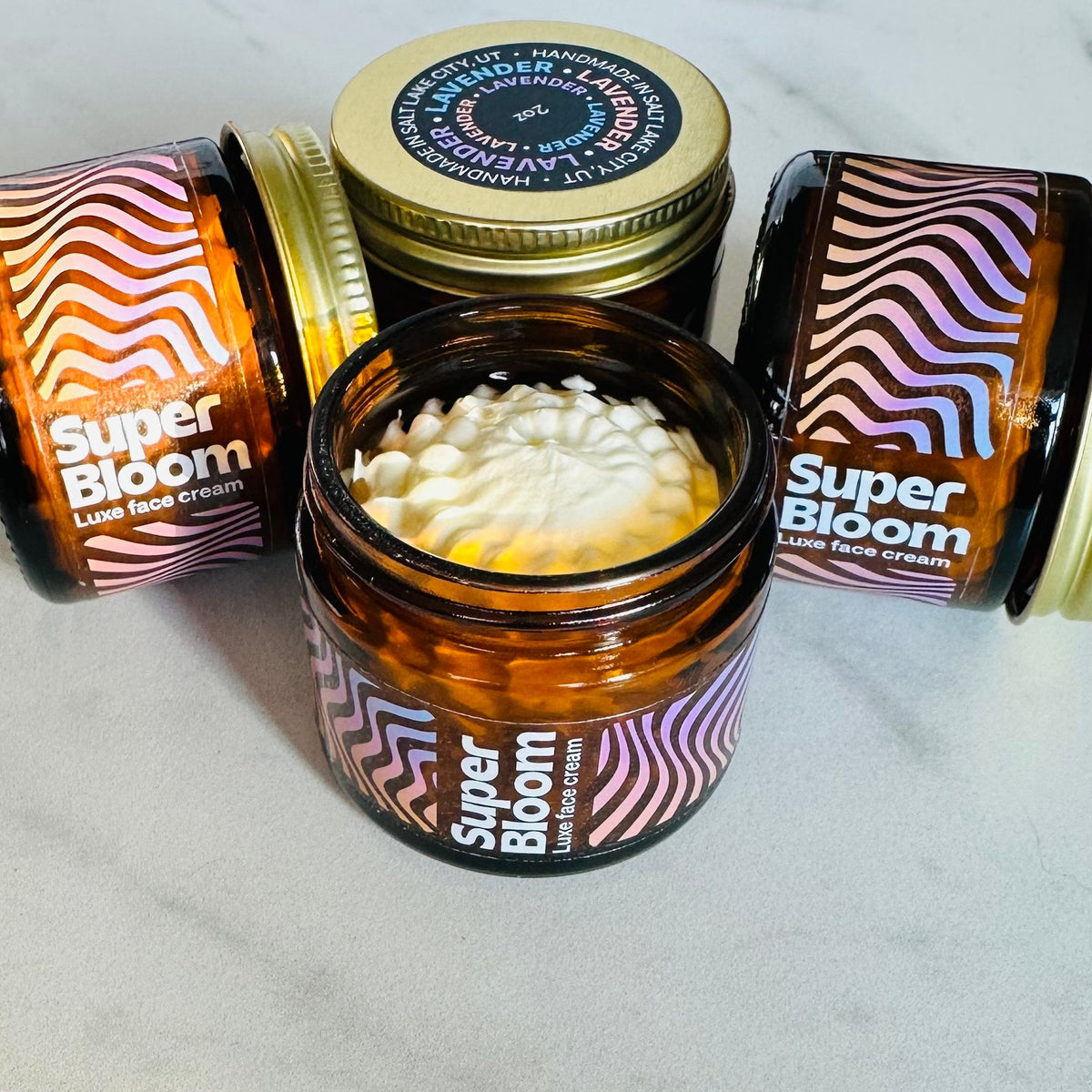 A collection of four small glass jars of handmade "Super Bloom" luxe face cream sits on a marble surface, with one open jar in the foreground revealing a fluffy white whipped cream texture inside, while the others are closed with amber glass and colorful wavy labels in shades of pink, purple, and white. Three jars have gold or brass lids, one featuring a navy blue sticker with "Lavender" text and "Handmade in Salt Lake City, UT" details, all arranged casually with some lids removed and placed nearby.