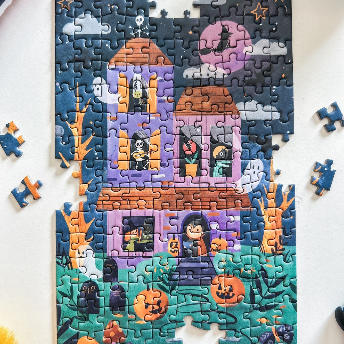 Mini Puzzle depicting a cute spooky mansion at night with jack-o-lanterns, ghosts, skeletons, zombies and witches. Pictured against a white back ground.