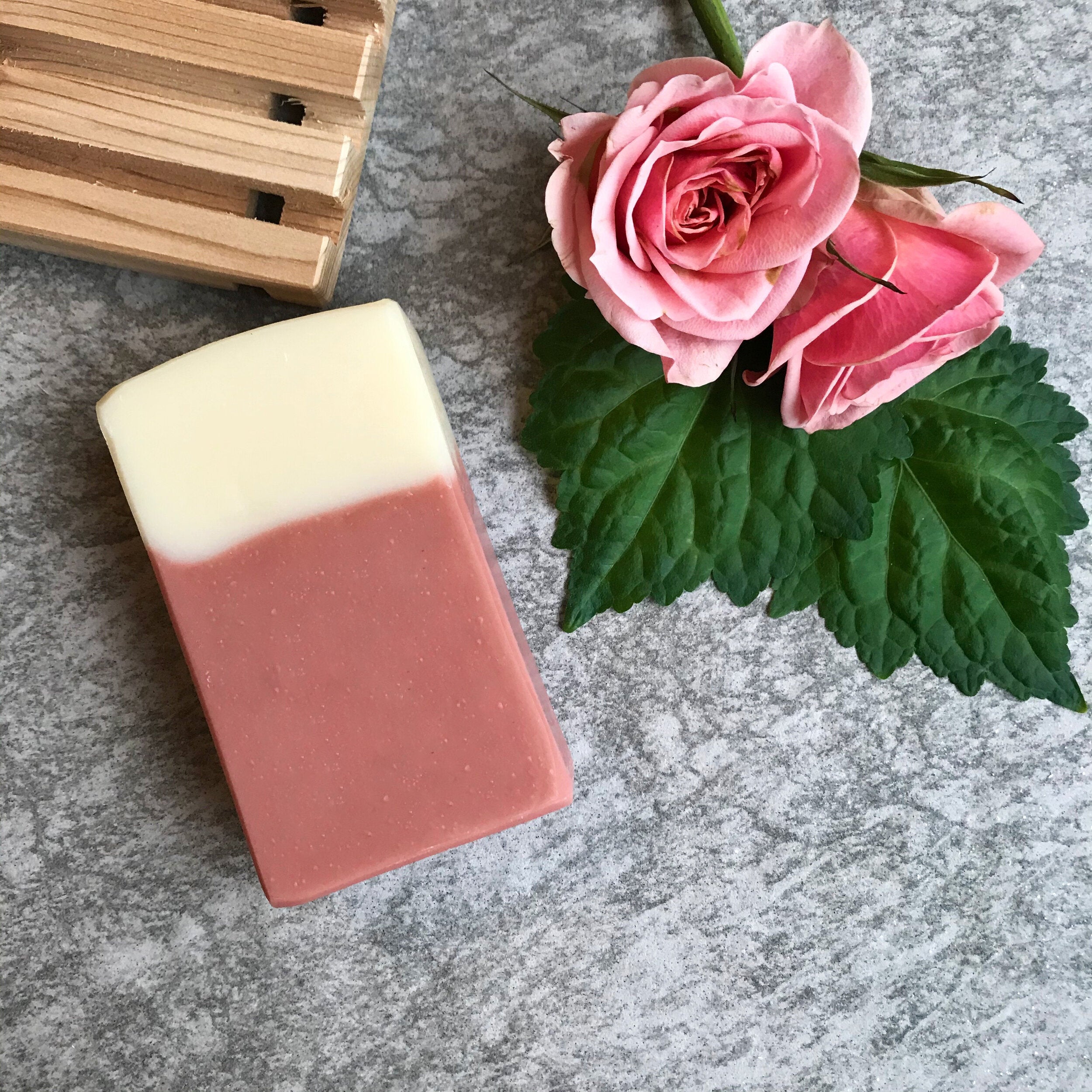Bar of soap with a gradient from white to pink, next to two pink roses on a gray surface.
