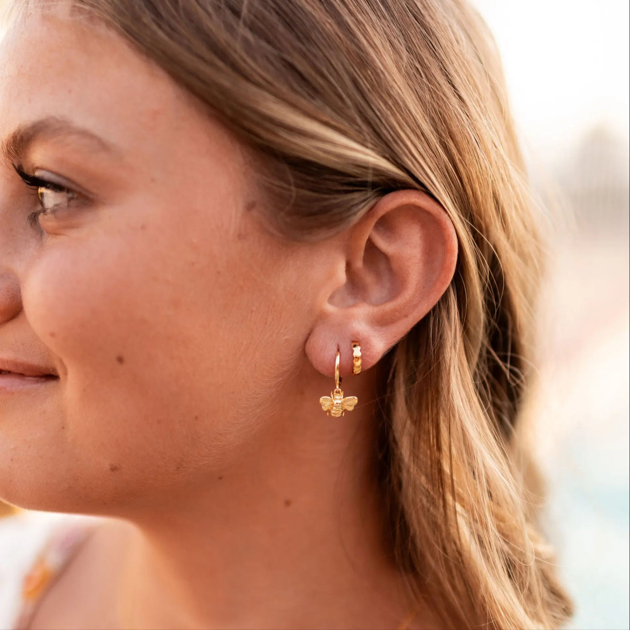 The left side profile of a model who is looking off-camera with two earrings in her ear. One is a honeybee and one is the earrings featured in this product listing, the honeycomb huggie. Both earrings are gold.