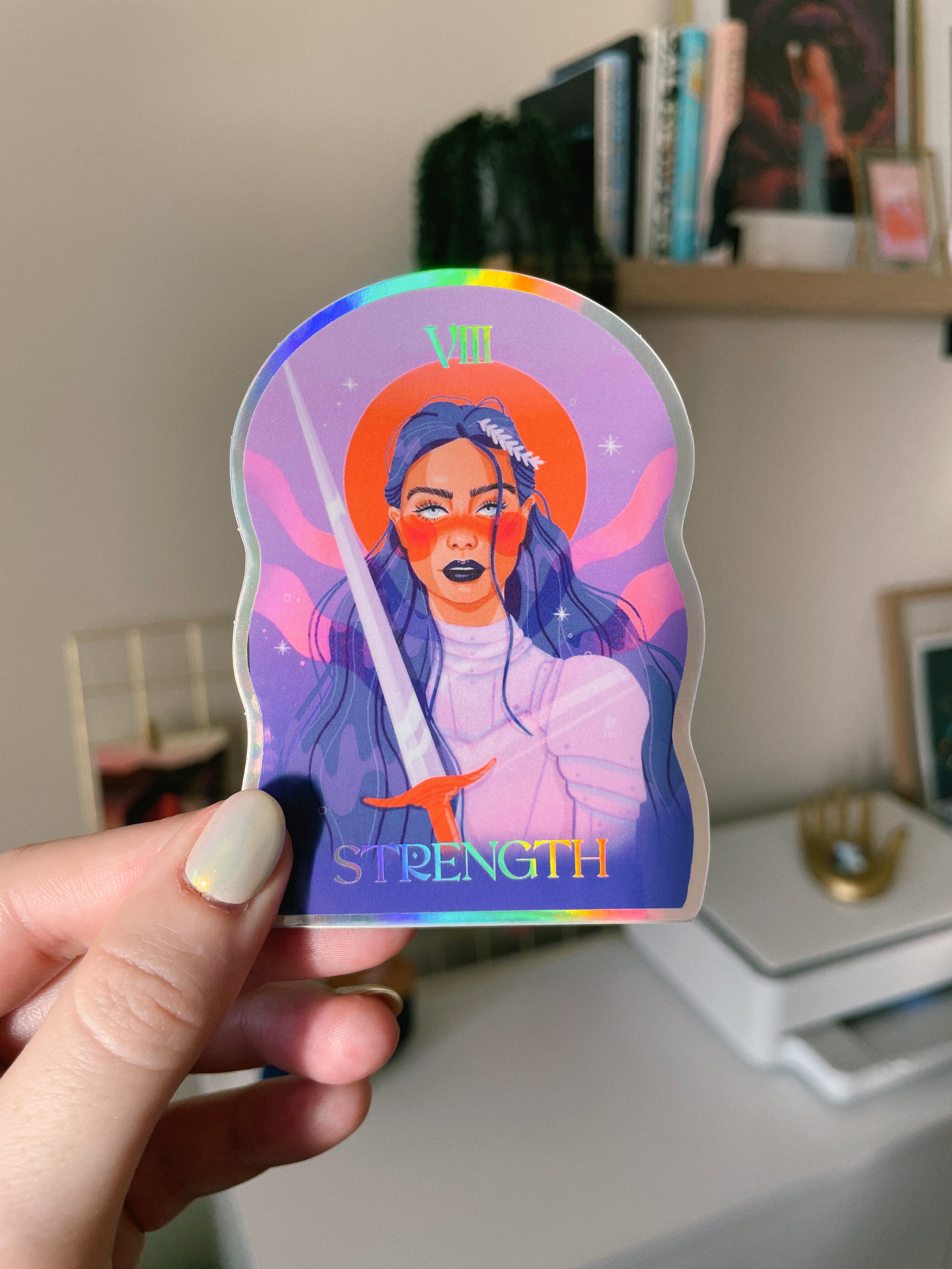 Strength sticker with holographic effects. The woman is holding a sword and she has long, blue-black hair and is wearing silver armor. The word "strength" is at the bottom of the sticker in holographic font.