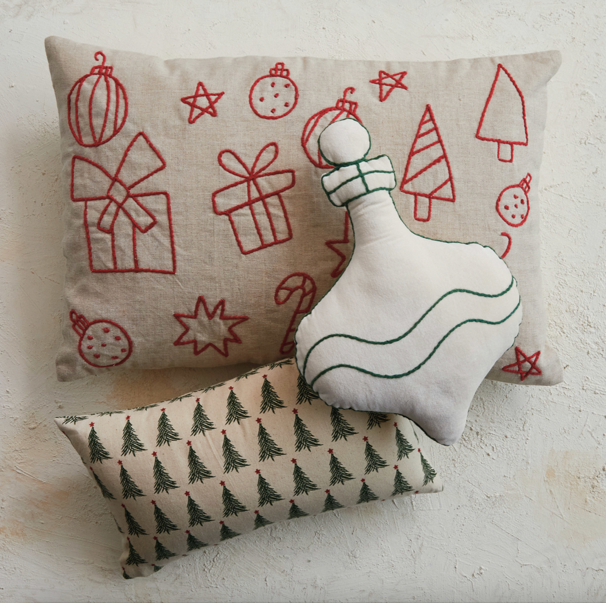 Three Christmas-themed decorative pillows arranged on a textured off-white wall: a large beige pillow with red embroidered holiday motifs, a smaller beige pillow with green tree prints, and a white ornament-shaped pillow with green stitched details.