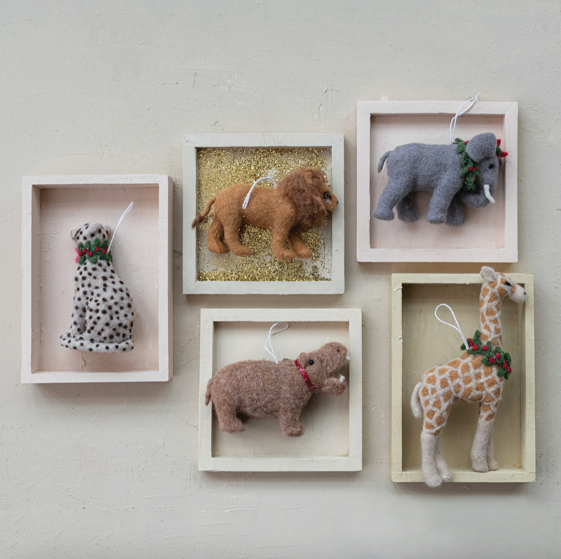 Five felt animal ornaments displayed in small framed backdrops on a light wall: a dalmatian, lion, elephant, hippo, and giraffe, each with a green garland detail.