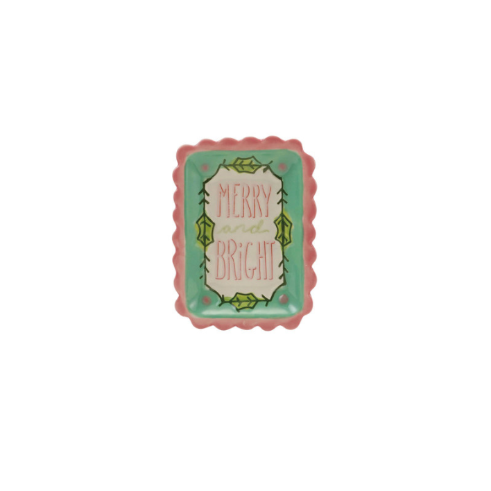 A small rectangular ceramic dish with a scalloped pink border and a turquoise center, featuring the words “MERRY and BRIGHT” in pink script surrounded by green leafy illustrations. The dish is photographed against a plain, textured off-white wall.