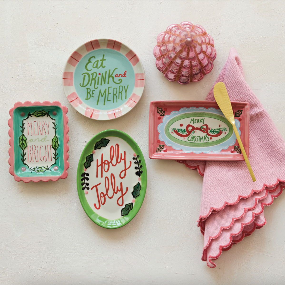 A styled flat-lay of a holiday table setting featuring a pink and white striped plate that reads 'Eat DRINK and BE MERRY', a green oval plate reading 'Holly Jolly', a pink rectangular dish with 'MERRY CHRISTMAS', a pink napkin, a yellow spoon, and a pink textured ornament on a textured off-white background.