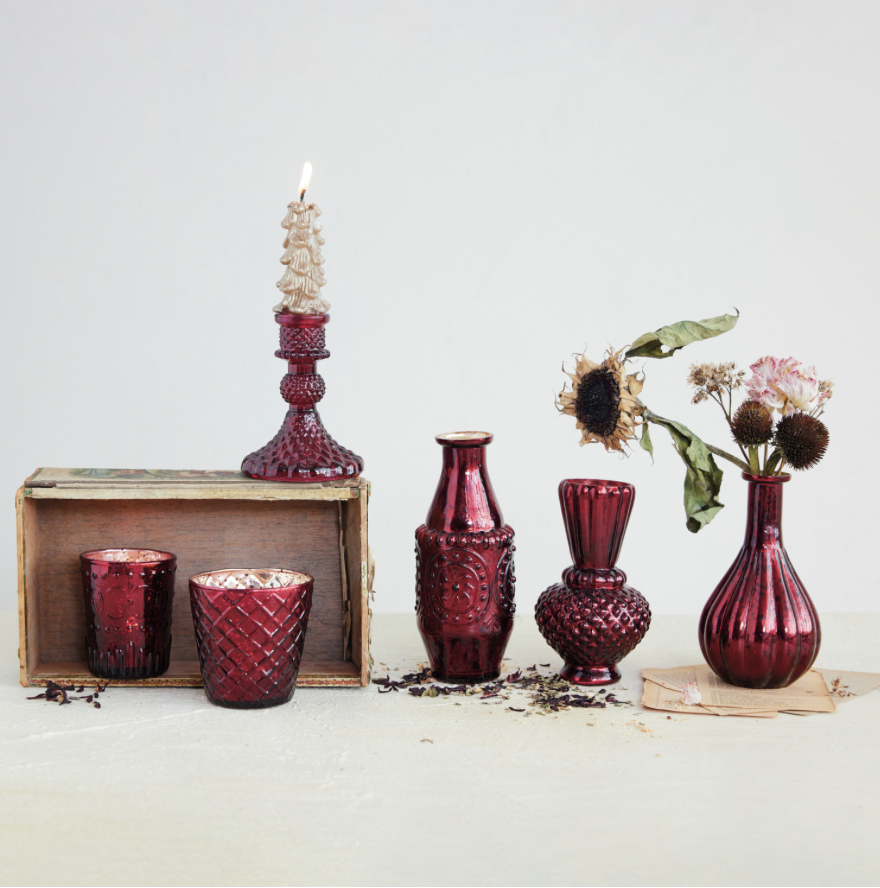 A coordinated set of deep burgundy glass vases and candle holders arranged on a light surface against a pale gray background. The set includes two small textured cups, a tall candlestick holder with a lit cream candle, a narrow bottle, a rounded vase, a small bulb-shaped vase, and dried flowers arranged in one of the vases.