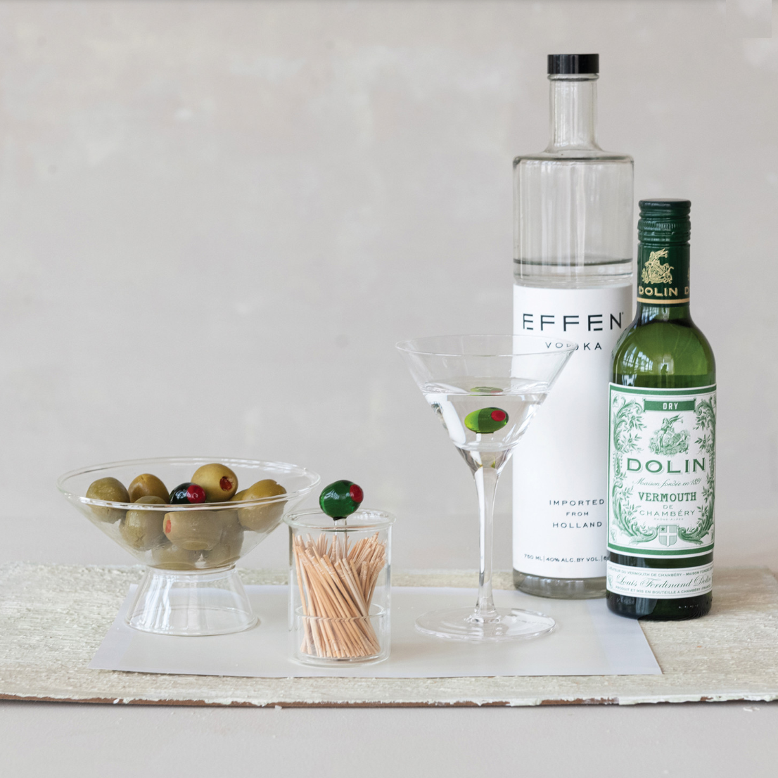 Bottles of vodka and vermouth with a glass and olives on a wooden surface.