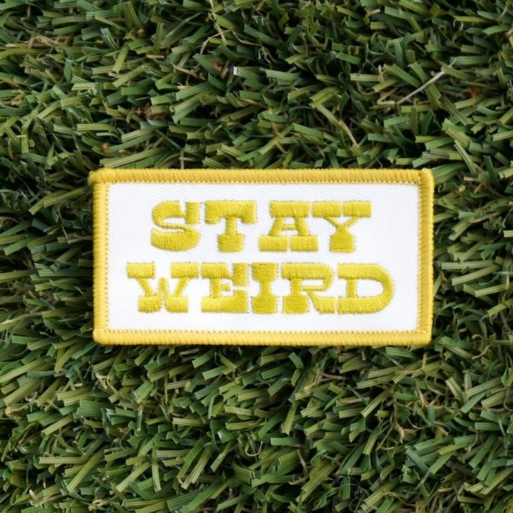 Iron-on rectangular patch with a white background and yellow words that say "Stay Weird".