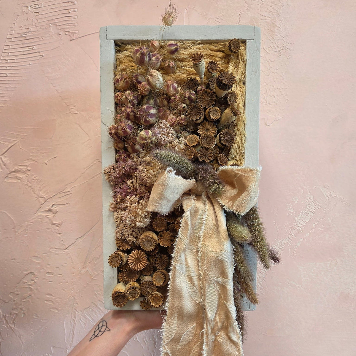 An arrangement of dried flora and fauna in a light gray 8 x 14 wooden frame. Hues of gold, pink, and light green fill the space, and there is a cream colored ribbon beautifully adorned in the lower right quadrant.