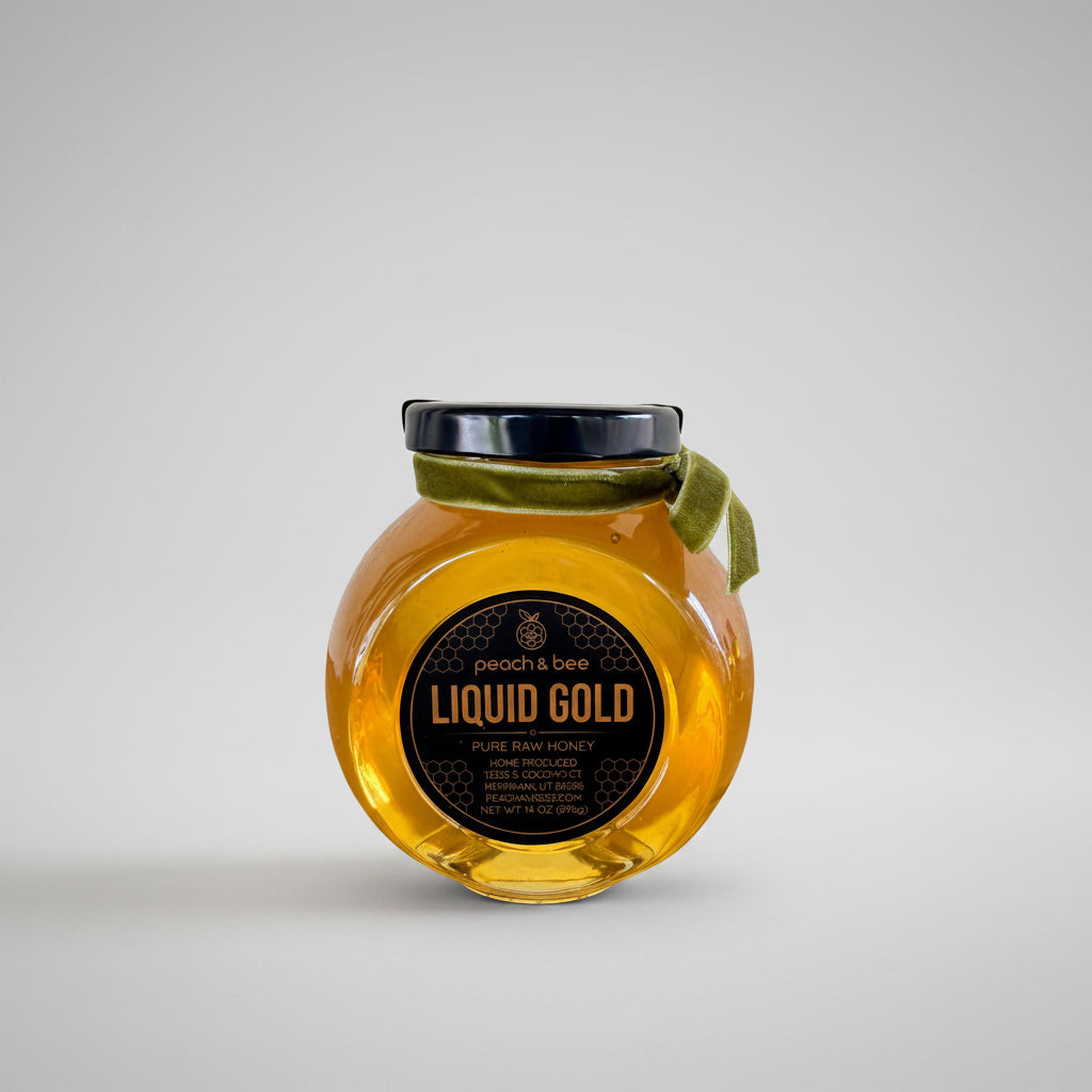 A glass jar of peach & bee Liquid Gold pure raw honey with a black lid, a green ribbon bow, and a black honeycomb-patterned label, shown on a white background.