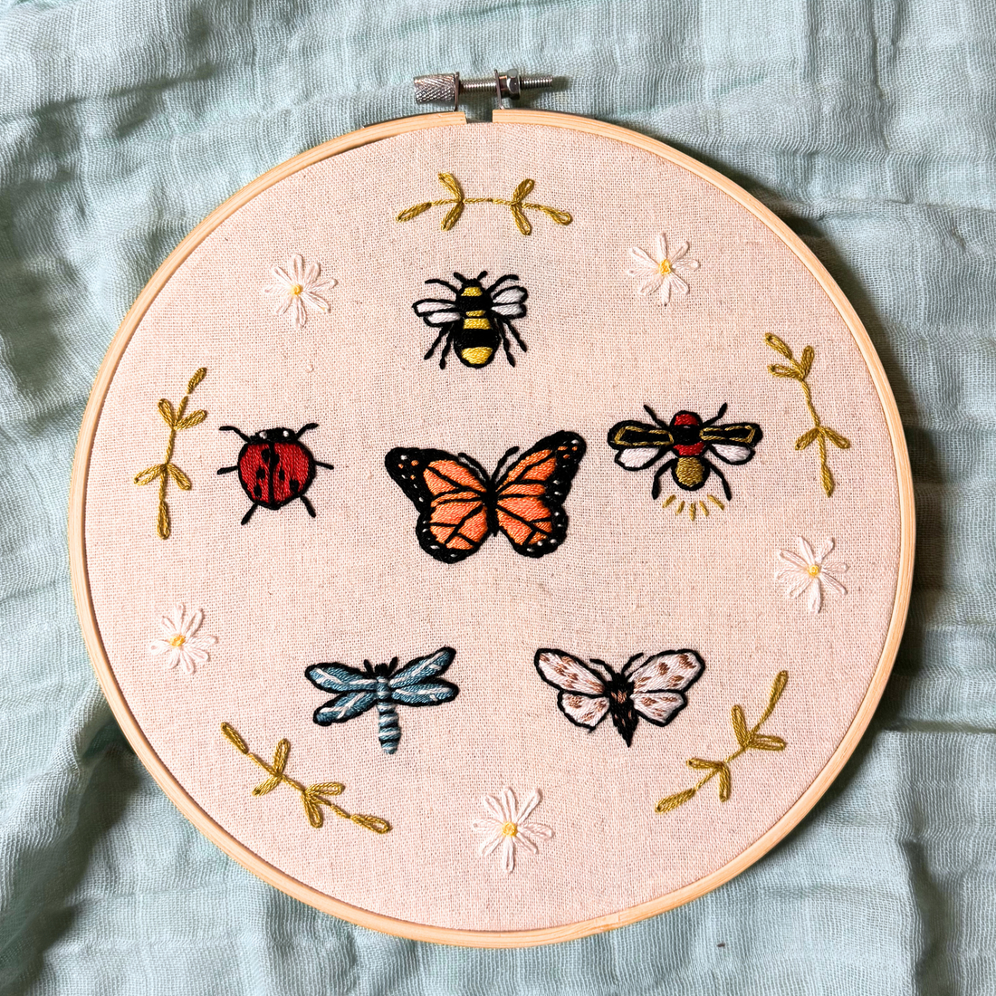 8 inch embroidery hoop featuring a wide variety of bugs (bumble bee, ladybug, dragonfly, beetle, butterfly, and moth) surrounded by flowers and greenery