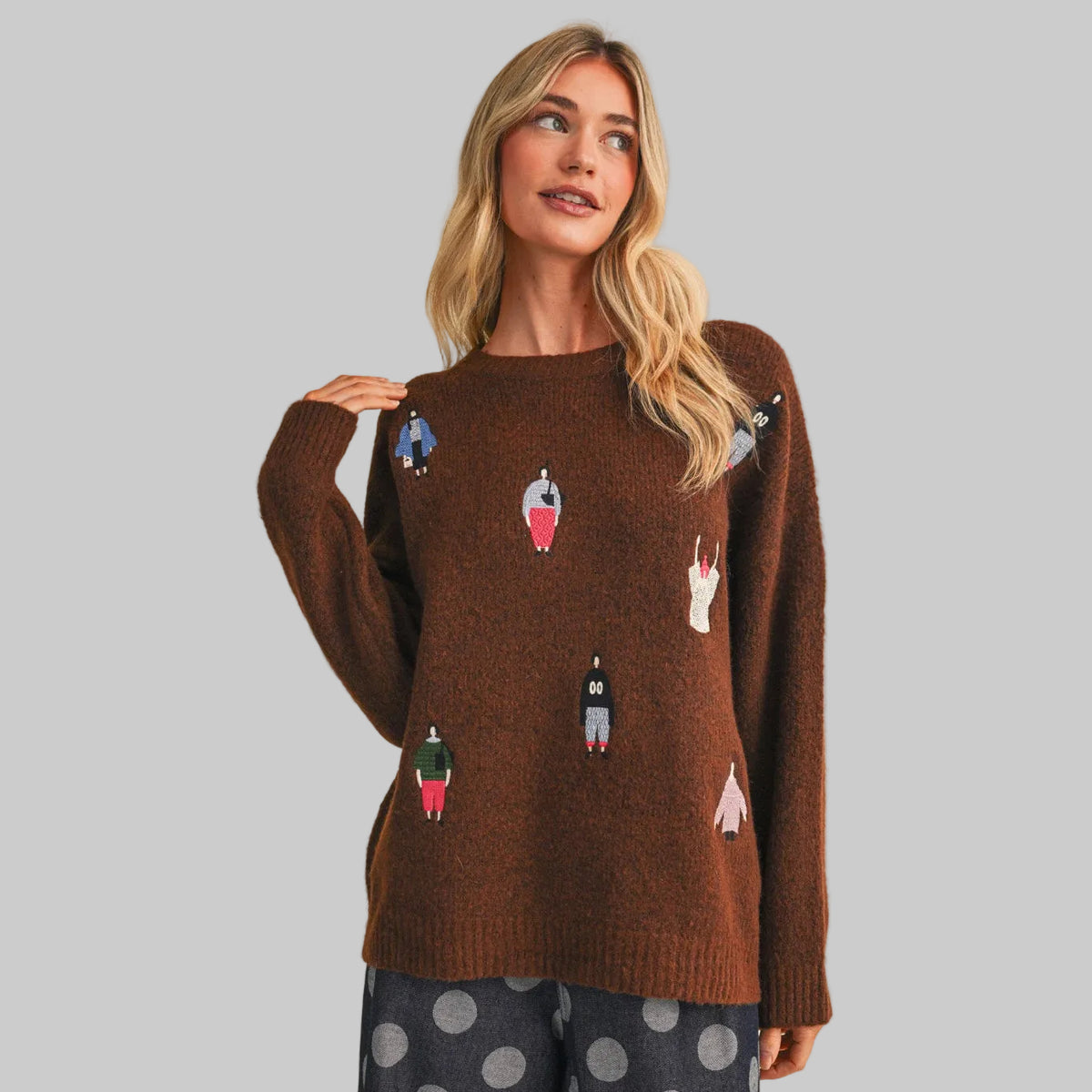 A woman wearing a brown knit sweater with small colorful women dreesed in fun clothing patches, paired with gray polka-dot pants, against a plain beige studio background.