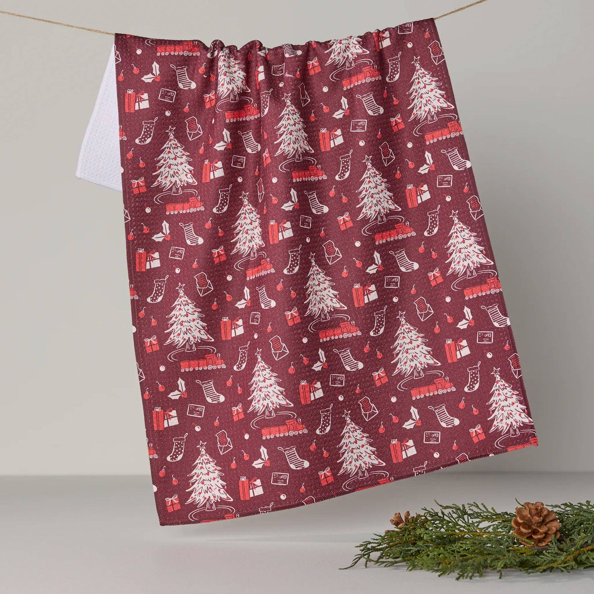 A dark red kitchen towel with a white and red Christmas pattern, hanging from a string on a neutral background with a pine branch prop.