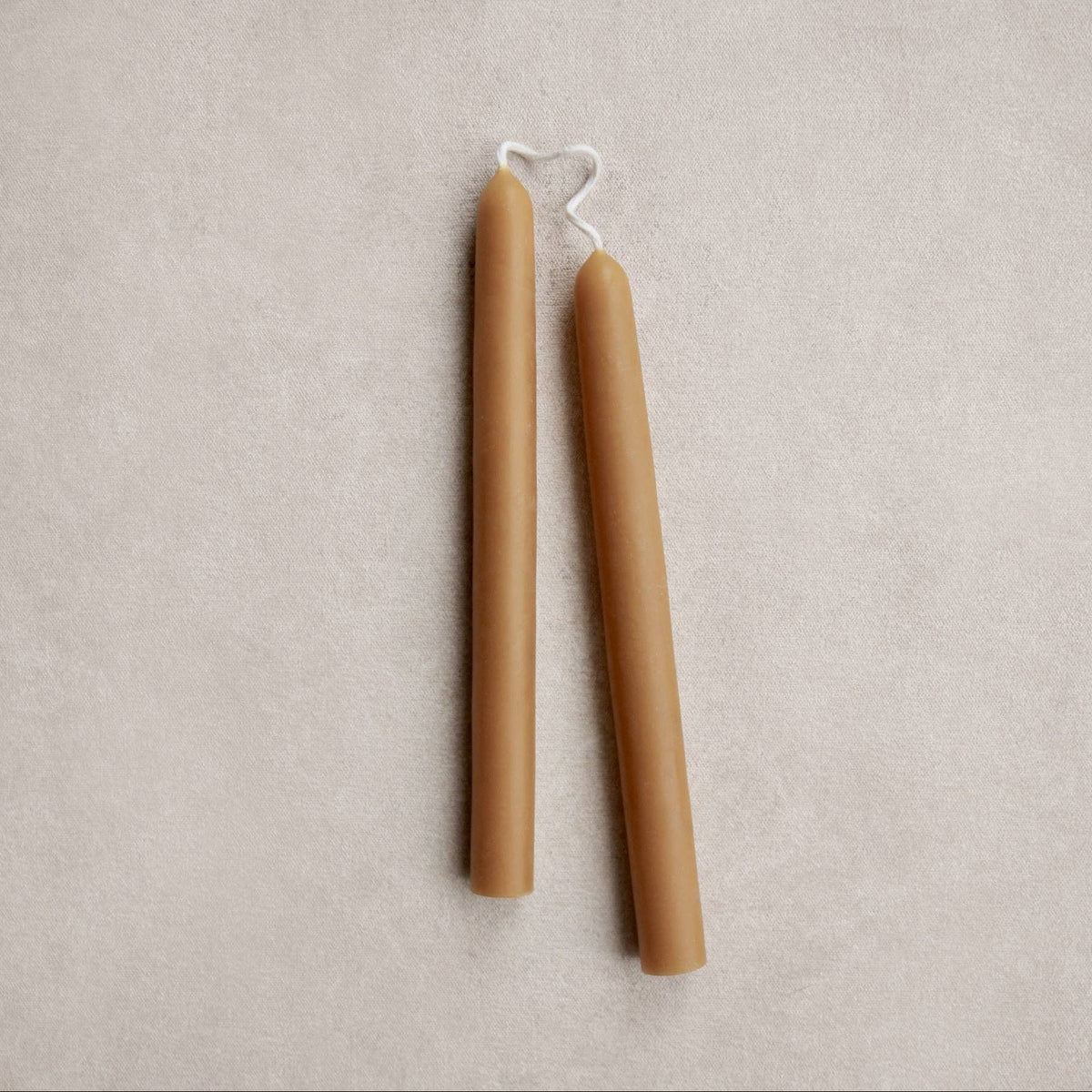 Two unpainted taper candles in a warm ochre color, standing upright on a textured beige surface.