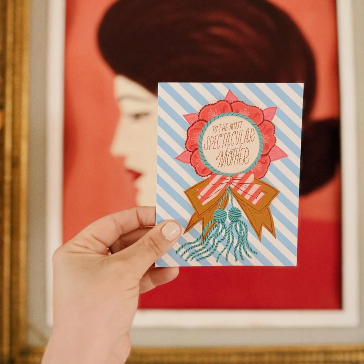Hand holding a card with decorative design in front of a framed portrait.