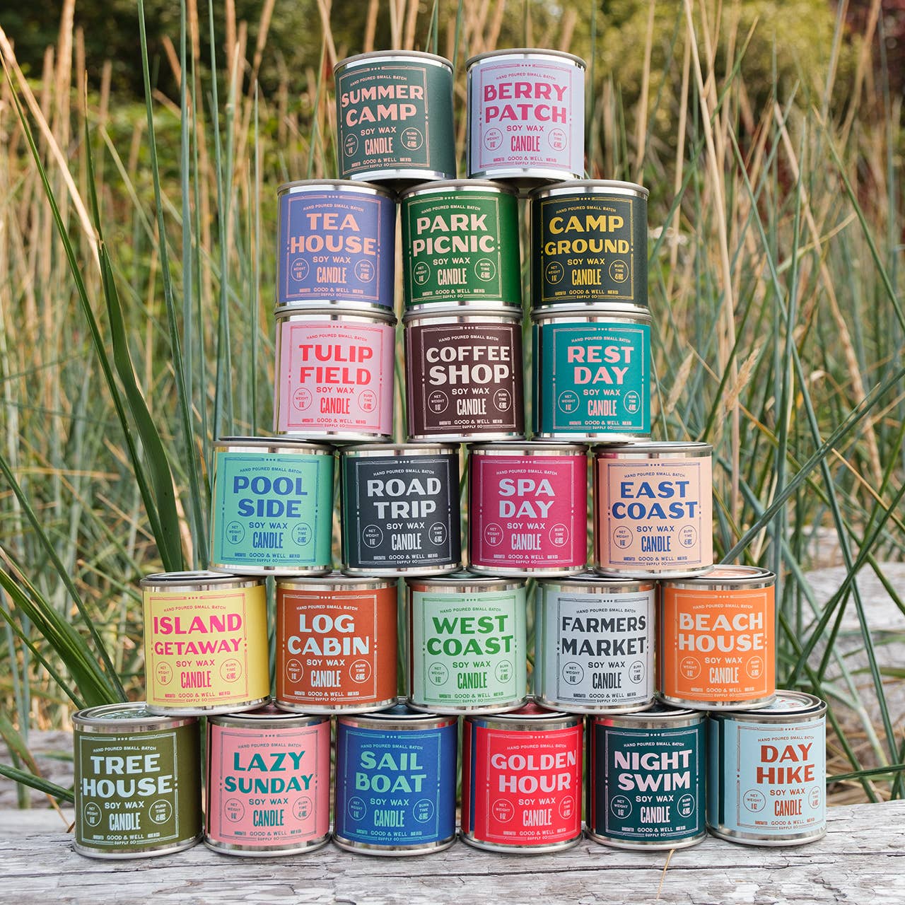 Stack of candles with various scents in cans on a wooden surface with grass in the background