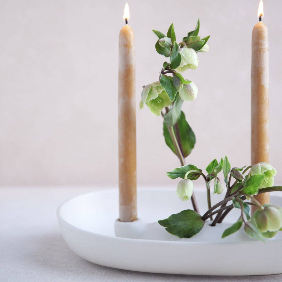 A white ceramic candle holder with two lit beige taper candles and green hollyhock branches, set against a soft, neutral background.